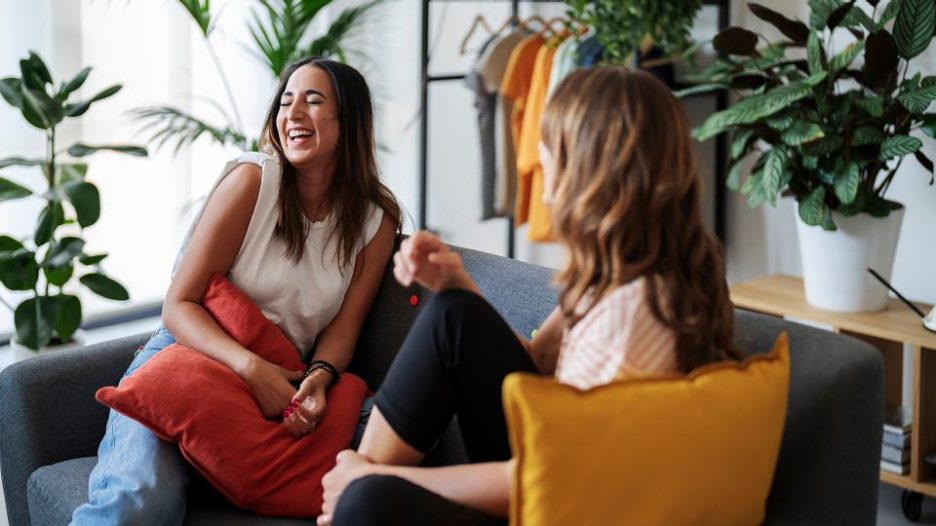 Young female in casual clothes smiling and sharing social media data on cellphone with Arab girlfriend while resting on couch in living room at home.