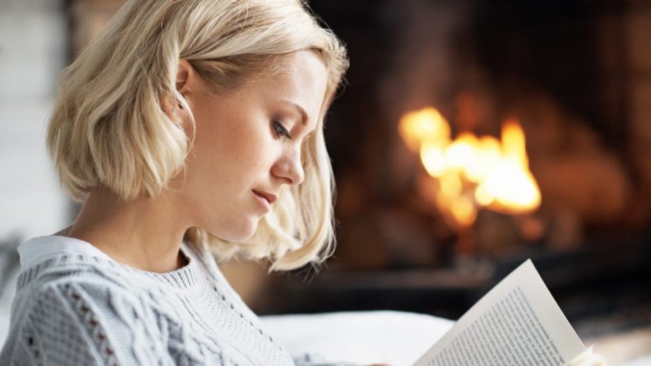 woman in a pale blue jumper reads a book by a fireplace