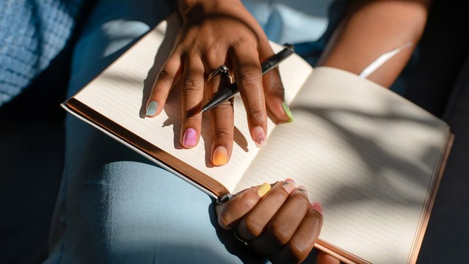 Close up of the hands of a woman holding a blank notebook and a pen. The nails have colorful and stylish nail art.