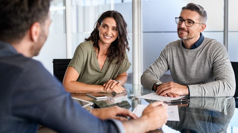 Smiling mature couple meeting with bank manager for investment. Beautiful mid adult woman with husband listening to businessman during meeting in conference room in modern office. Happy middle aged couple meeting loan advisor to buy a new home.